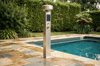 Douche solaire en inox installée en bord de piscine avec dallage en pierre naturelle et haie verte en arrière-plan