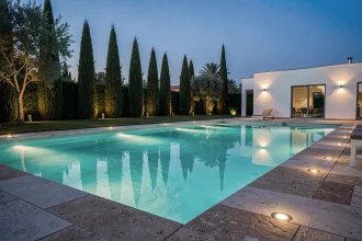 Piscine contemporaine illuminée le soir avec des LED sous-marines turquoise et des spots encastrés au sol autour de la margelle en pierre naturelle