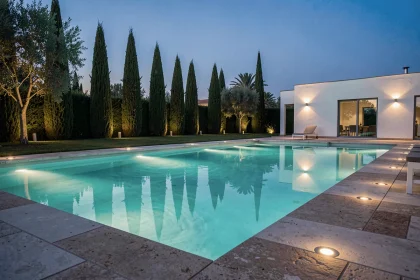 Piscine contemporaine illuminée le soir avec des LED sous-marines turquoise et des spots encastrés au sol autour de la margelle en pierre naturelle